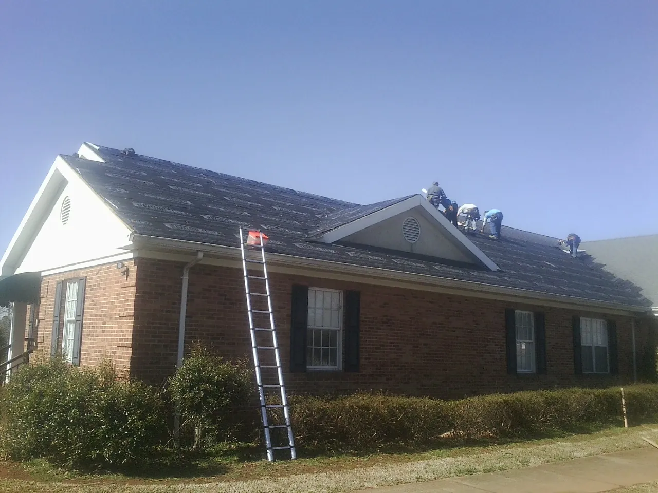 Crew tearing shingles off a brick ranch roof with a ladder set — Metal Roof Repair work by our crew in Nashotah