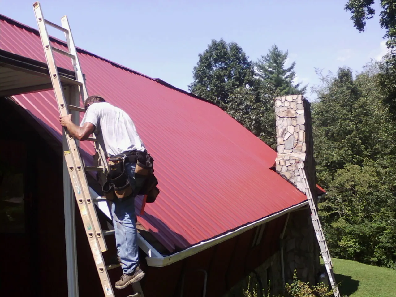 Roofer on a ladder beside a red metal roof and stone chimney — Soffit & Fascia Repair by Roofing Brothers in Nashotah